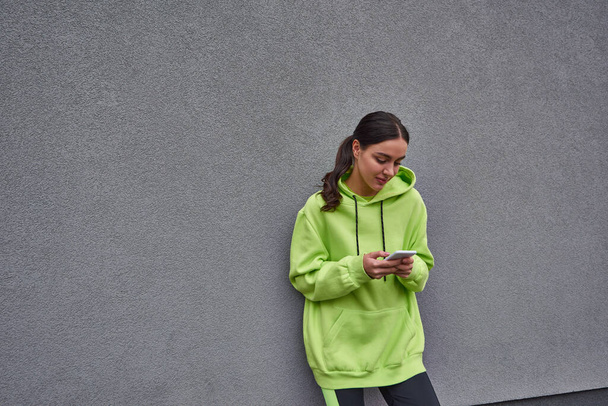 jonge vrouw in kalk kleur hoodie en legging messaging op smartphone in de buurt van grijs beton muur - Foto, afbeelding