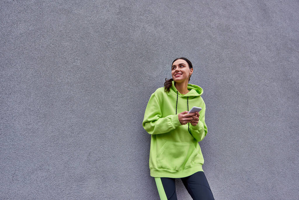 Limon rengi kapüşonlu ve taytlı mutlu kadın gri beton duvarın yanındaki akıllı telefondan mesajlaşıyor. - Fotoğraf, Görsel