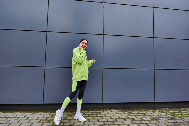 happy woman in lime color hoodie and leggings talking on smartphone near grey wall outdoors - Photo, Image