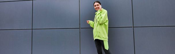 happy woman in lime color hoodie and leggings talking on smartphone near grey wall outdoors, banner - Photo, Image