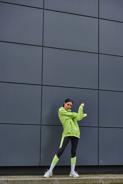 bonita deportista con capucha y polainas calentándose cerca de la pared gris al aire libre, aire fresco y deporte - Foto, Imagen