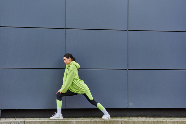 νεαρή αθλήτρια με κουκούλα και κολάν που τεντώνουν τα πόδια κοντά σε γκρίζο τοίχο σε εξωτερικούς χώρους, ενέργεια και αθλητισμό - Φωτογραφία, εικόνα