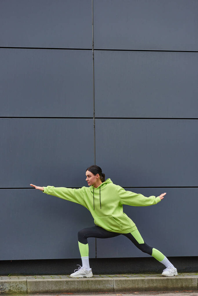 νεαρή αθλήτρια σε αθλητικά ρούχα με απλωμένα χέρια που τεντώνουν τα πόδια κοντά σε γκρι τοίχο σε εξωτερικούς χώρους - Φωτογραφία, εικόνα