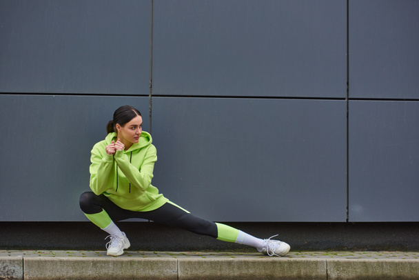 jeune sportive en sweat à capuche et leggings faisant des fentes près du mur gris à l'extérieur, l'énergie et le sport - Photo, image