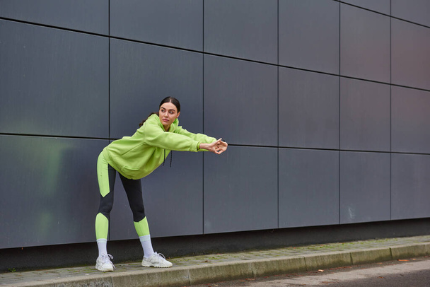 atlétikai nő kapucnis pulóverben és lábszárvédőben bemelegítés a szürke fal közelében szabadban, motiváció és sport - Fotó, kép