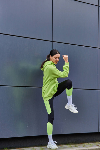 atletyczna biegaczka w kapturze i legginsach rozgrzewająca się w pobliżu szarej ściany na zewnątrz, motywacja - Zdjęcie, obraz