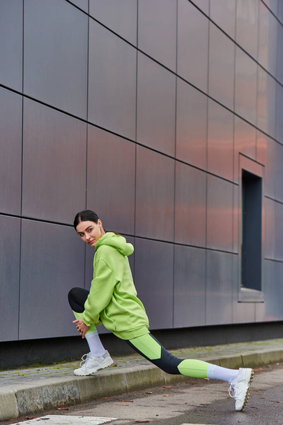 pretty female runner in hoodie and leggings doing lunges near grey wall outdoors, motivation - Photo, Image