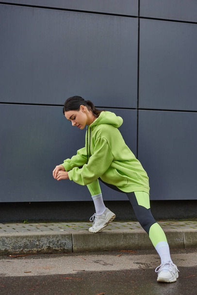 athletic woman in lime color hoodie and leggings stretching near grey wall outdoors, motivation - Photo, Image