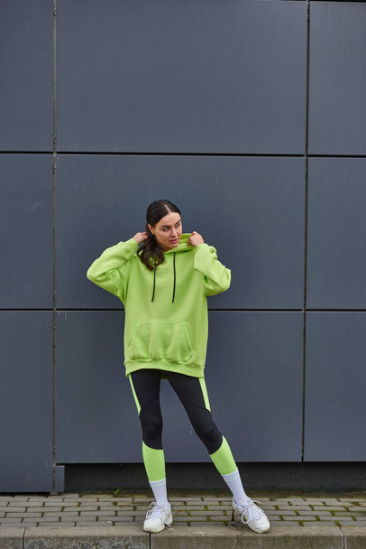 attraente sportiva con cappuccio color lime e leggings in piedi vicino al muro grigio all'aperto, sport - Foto, immagini