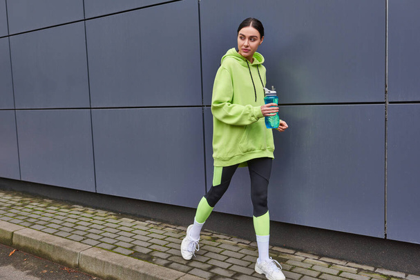 mooie sportvrouw in hoodie en legging wandelen met fles water in de buurt van grijze muur buiten - Foto, afbeelding