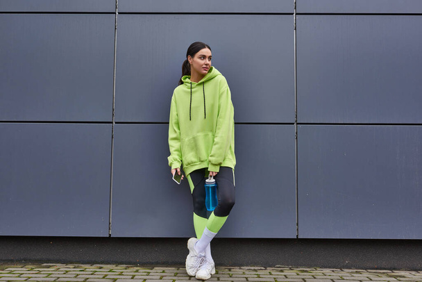 schöne Sportlerin im Kapuzenpullover mit Wasserflasche und Smartphone in der Nähe der grauen Wand im Freien - Foto, Bild