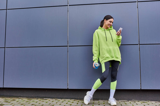 χαρούμενη αθλήτρια με κουκούλα που κρατάει μπουκάλι νερό και χρησιμοποιεί smartphone κοντά σε γκρίζο τοίχο σε εξωτερικούς χώρους - Φωτογραφία, εικόνα