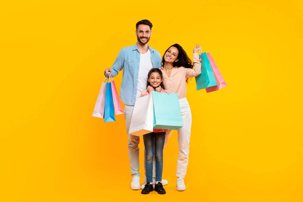 Счастливая семья держит сумки с покупками и улыбается, родители и их дочь позируют с красочными покупателями бумаги, стоя на желтом фоне студии. Реклама сезона продаж - Фото, изображение