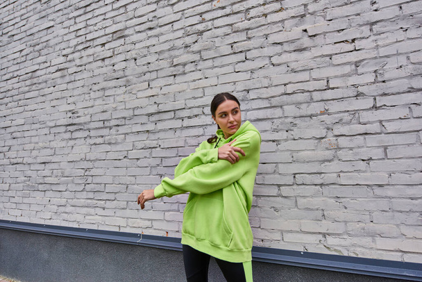 attraktive junge Sportlerin in lindfarbenem Kapuzenpulli und Leggings, die sich in der Nähe der grauen Wand im Freien ausstrecken - Foto, Bild