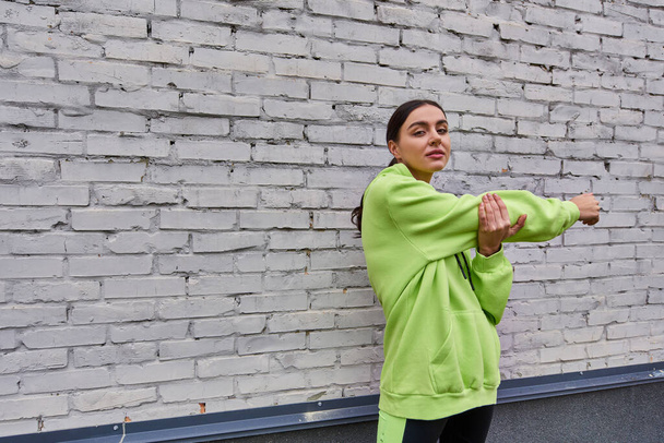 παρακινούμενη νεαρή αθλήτρια με lime χρώμα φούτερ και κολάν που εκτείνεται κοντά σε γκρίζο τοίχο σε εξωτερικούς χώρους - Φωτογραφία, εικόνα