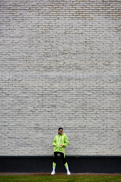 レンガの壁の近くのポケットに手で立っているライムカラーパーカーとレギンスの美しいスポーツウーマン - 写真・画像