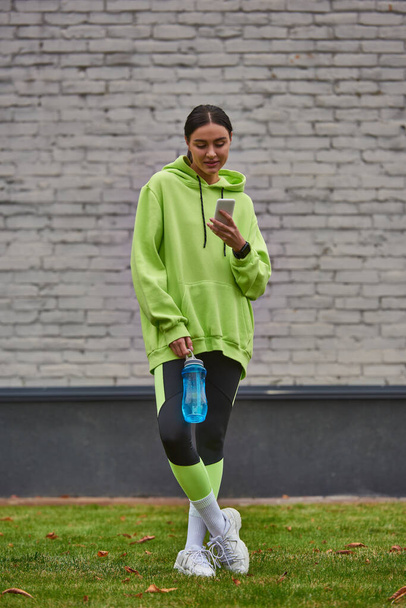 brunette sportswoman in hoodie and leggings using smartphone and standing with bottle with water - Foto, imagen