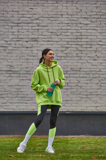 веселая спортсменка в толстовке и леггинсах, держащая смартфон и стоящая с бутылкой с водой - Фото, изображение