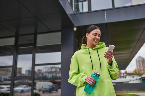 χαρούμενη αθλήτρια με ζεστή κουκούλα χρησιμοποιώντας smartphone και στέκεται με μπουκάλι με νερό σε εξωτερικούς χώρους - Φωτογραφία, εικόνα