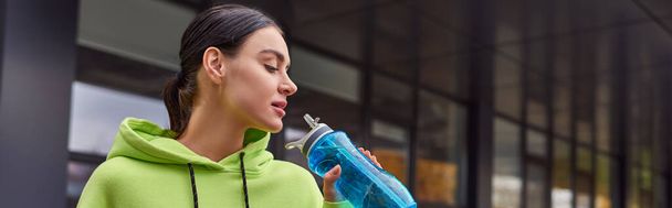 vrij sportvrouw in limoen kleur hoodie drinkwater uit fles na training, horizontale banner - Foto, afbeelding