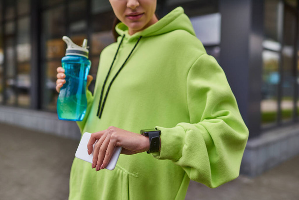 uprawiana sportowiec trzymając smartfona i butelkę podczas sprawdzania fitness tracker po treningu - Zdjęcie, obraz