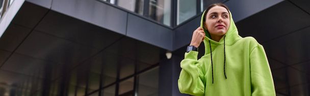 banner of young sportswoman with hood on head standing near modern building, lime color hoodie - Photo, Image