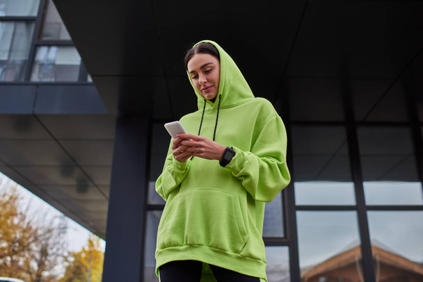 Kafasında kapüşonlu genç esmer kadın modern binanın yanında akıllı telefon kullanıyor, sokak tarzı. - Fotoğraf, Görsel