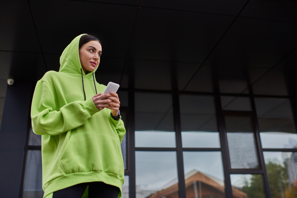 近代的な建物の近くのスマートフォンを使用して頭にフードを持つ若いブルネットの女性,ストリートスタイルの外観 - 写真・画像