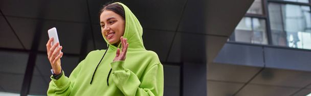 mujer joven feliz en sudadera con capucha saludando la mano mientras tiene videollamada en el teléfono inteligente cerca del edificio, pancarta - Foto, imagen