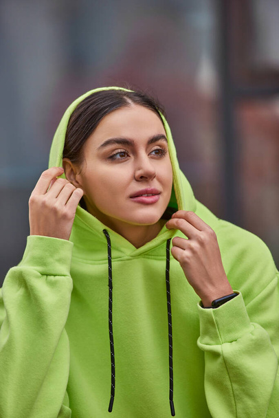 brunette young woman in lime color hoodie wearing hood on head and looking away outdoors - Photo, Image