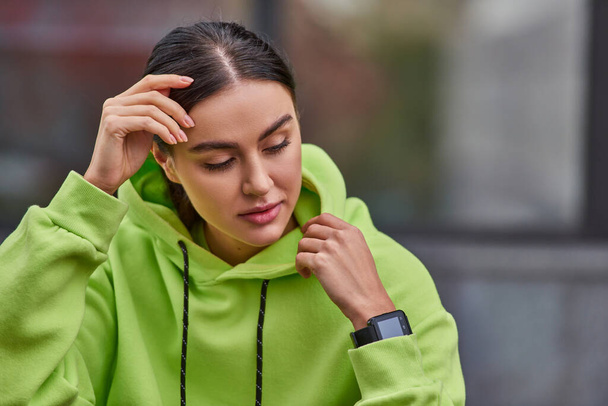 hübsche junge Frau in lindfarbenem Kapuzenpulli posiert mit smarter Uhr am Handgelenk und schaut weg - Foto, Bild