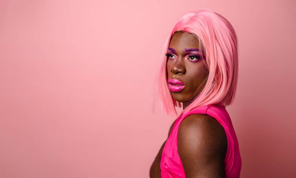 Νεαρό αυτο-εξασφαλισμένο αφροαμερικάνικο τρανσέξουαλ μοντέλο φορώντας ροζ περούκα και μακιγιάζ αγγίζοντας τον ώμο ενώ στέκεται με το χέρι στη μέση και κοιτάζοντας κενό χώρο για διαφήμιση - Φωτογραφία, εικόνα