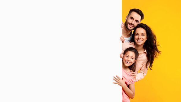 Oferta familiar. Emocionados padres europeos y su hija asomándose de la cartelera blanca en blanco con espacio libre para su anuncio y diseño, pancarta - Foto, Imagen