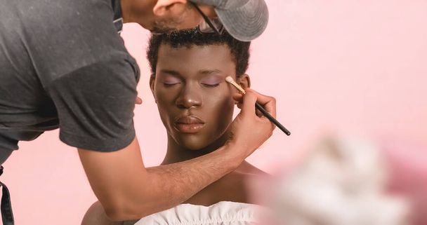 Cosecha artista de maquillaje concentrado aplicando sombra de ojos púrpura en el párpado de drag queen afroamericano sentado con los ojos cerrados sobre fondo rosa - Foto, imagen