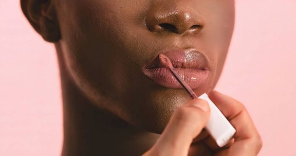 Primo piano di coltura visagiste irriconoscibile applicando lucidalabbra con pennello mentre fa il trucco sulla drag queen afroamericana con pelle liscia perfetta su sfondo rosa - Foto, immagini