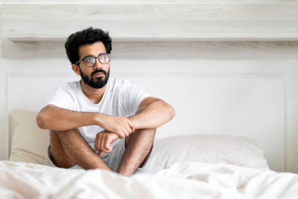 メランコリーコンセプト。 若い高価なインドの男はベッドに座って離れて見て,ライトベッドルームでリラックスしながら何かについて考える東部の男,苦しみの季節のうつ病や不快な - 写真・画像