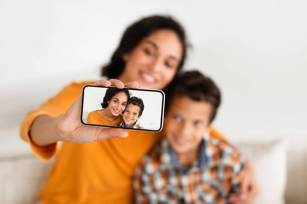 Bonne maman et fils enfant utilisant Smartphone faisant Selfie assis sur le canapé à la maison, foyer sélectif sur la photo sur l'écran de téléphone portable tandis que la mère étreint son enfant. Profondeur peu profonde. Médias sociaux, Gadgets - Photo, image