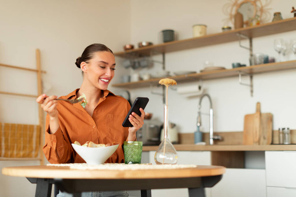 Веселая дама ест вкусный доставленный салат и с помощью мобильного телефона, сидя за столом на кухне во время ужина, женщина пишет отзыв о доставке и ресторанного обслуживания, копировать пространство - Фото, изображение