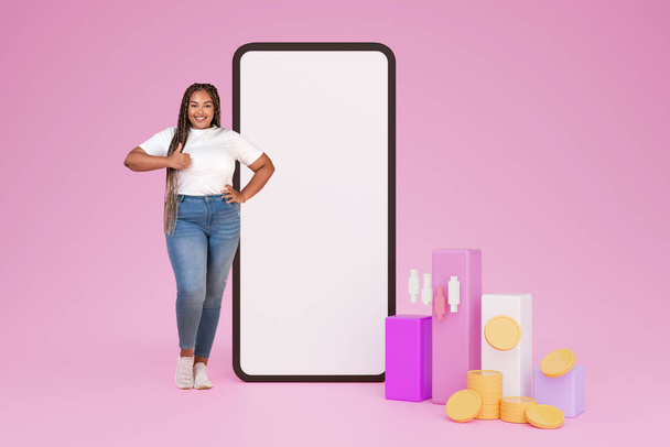 Šťastný baculatý mladý africký americká žena obchodník doporučující obchodování mobilní aplikace, stojící vedle velkého smartphonu s bílou prázdnou obrazovkou, mockup, akcie grafy a mince, ukazující palec nahoru, koláž - Fotografie, Obrázek