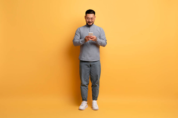 Young Smiling Asian Man With Smartphone In Hands Standing On Yellow Studio Background, Happy Millennial Guy Wearing Eyeglasses Using Mobile Phone, Browsing New App Or Messaging, Copy Space - Photo, Image