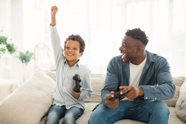 Videogaming Plezier. Joyful Black Daddy en zijn zoon winnen Online Video Game, vieren samen de overwinning, Kid Boy schudden vuist in triomf zittend op de bank thuis. Digitaal entertainment - Foto, afbeelding