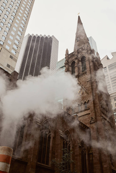 Blick auf die katholische Kirche aus rotem Backstein in der Nähe von Wolkenkratzern und Dampf auf der New Yorker City Street - Foto, Bild