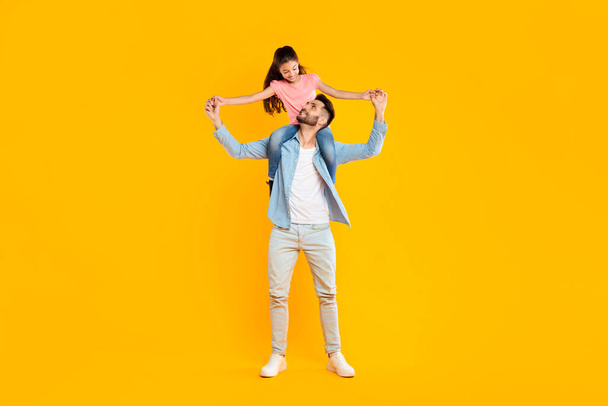 Ευτυχισμένες στιγμές. Ενθουσιασμένος άντρας να καβαλάει την κόρη του στους ώμους, να διασκεδάζει μαζί, πατέρας να κρατάει τα χέρια του παιδιού και να χαμογελά ο ένας στον άλλο σε κίτρινο φόντο, σε όλο το μήκος - Φωτογραφία, εικόνα