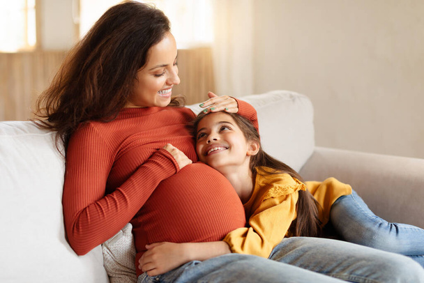 Zweite Schwangerschaft, Warten auf den kleinen Bruder. Mädchen berührt ihren Schwangerschaftsbauch und lächelt Mama zu Hause an. Erwartende Frau sitzt mit ihrer Tochter auf Couch im Wohnzimmer - Foto, Bild