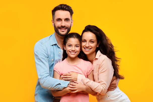Šťastní evropští rodiče objímají svou krásnou dceru a usmívají se na žluté pozadí, studiový záběr. Manželé objímání a pózování s dívkou - Fotografie, Obrázek