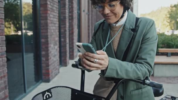 Tilt up shot of modern businesswoman with bicycle standing outdoors searching for address on map app on smartphone - Footage, Video