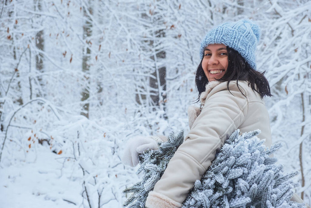 En el bosque nevado en medio del invierno, la mujer se para sosteniendo el árbol de Navidad nevado, sonriendo felizmente - Foto, imagen