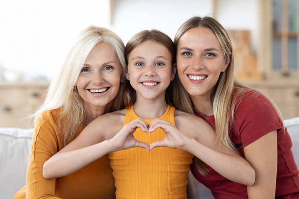 Glücklich schöne blonde kaukasische Mutter, Tochter und Enkelin posieren zusammen zu Hause. Nettes Teenie-Mädchen mit herzförmiger Geste und Lächeln in die Kamera mit ihrer Mutter, Großmutter - Foto, Bild