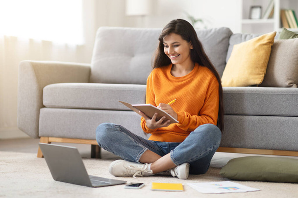 Beautiful cheerful millennial indian woman student have online lesson with tutor teacher, sitting on floor in living room, looking at laptop screen, taking notes in notepad, home interior, copy space - Photo, Image