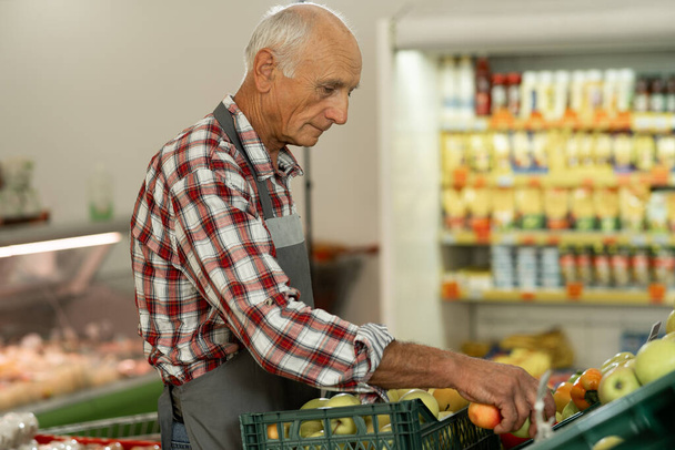 Egy idős férfi munkás gyümölcsöt tesz a zöldségosztályra az ökotermékek polcaira. Fénymásolási hely - Fotó, kép